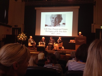 A panel of four seated people on stage with a person standing at a lecturn, with a slide showing on a screen above - a portrait of Anthony Ryle, smiling