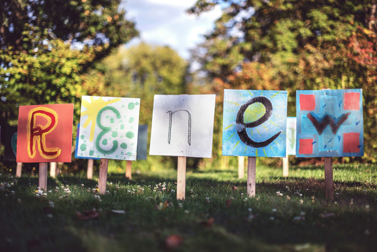 Five placards planted in a  grassy lawn with trees in the backgroun. They each display a letter and spell out R-e-n-e-w in colourful hand-painted images