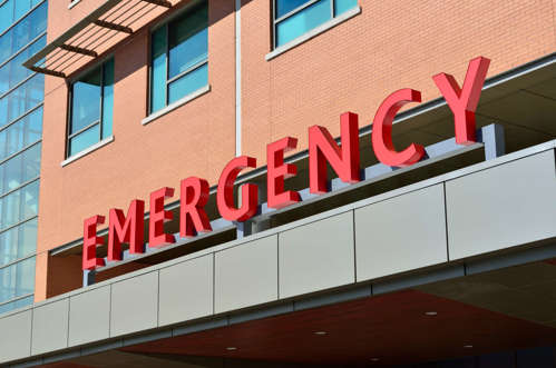 Red lettering signage above a grey hospital building canopy,  spelling the word EMERGENCY